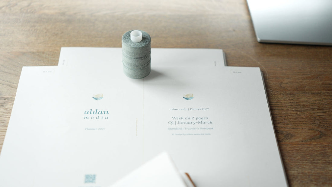 The image shows a spool of grey yarn standing upright on printed copies of our planner inserts. The printouts are our W2P, weekly, week on 2 pages for Traveler's Notebook in standard size in the colour Ming/Blue..